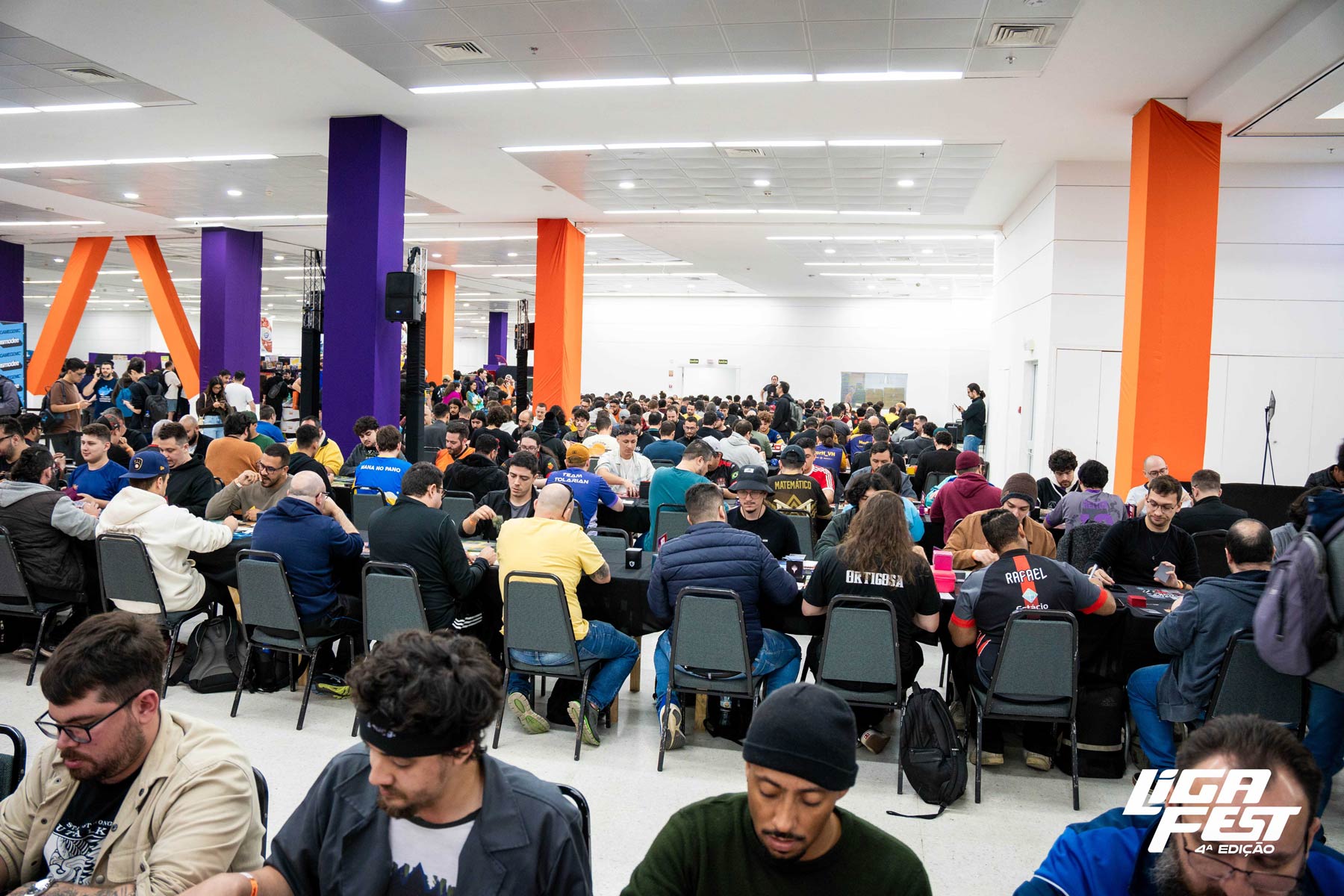 Salão da LigaFest lotado de jogadores em mesas de torneio, entre pilares roxos e laranja